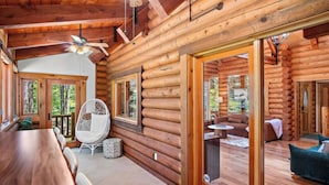 Une cabane en rondins avec un intérieur en bois, un coin repas avec une table et des chaises, un espace détente avec un canapé et un fauteuil, et une vue sur l’extérieur grâce à une porte vitrée.