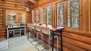 L’intérieur d’une cabane en rondins, avec une table à manger en bois, des tabourets de bar et de grandes fenêtres donnant sur la forêt.