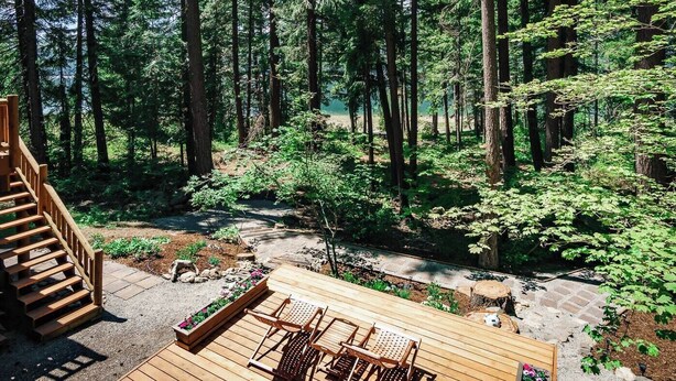 Une terrasse en bois avec des sièges extérieurs, entourée d’une zone boisée avec de grands arbres et un sentier bien dégagé.