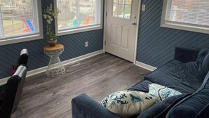 A room with dark blue walls, a white door, and two windows with blinds. There is a blue couch with patterned cushions, a small white table with a plant, and a decorative anchor on the wall.