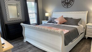 A bedroom with a white bed, dark curtains, a wall clock, and a mirror.