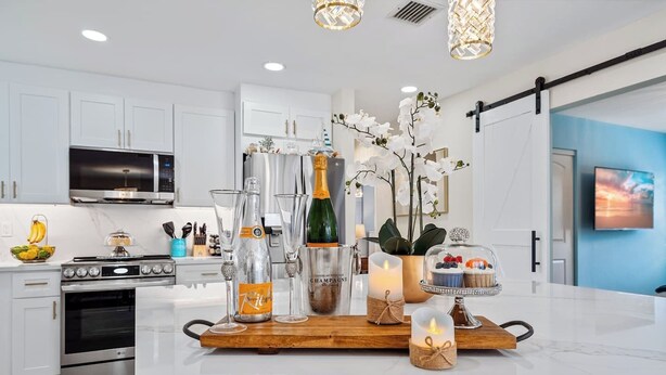 Une cuisine moderne avec des armoires blanches, une cuisinière en acier inoxydable et un grand îlot central doté d’un plan de travail en verre. L’îlot est agrémenté de divers objets de cuisine et éléments de décoration, notamment une bouteille de champagne, un vase avec des fleurs et un chandelier.