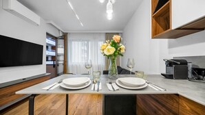 A modern dining area with a wooden table set for two, a vase of flowers, and a kitchen area with a built-in oven. - Warsaw Postępu Apartment - Parking, Balcony, A/C - by Rentujemy (Warszawa)