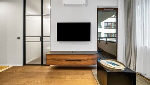 A modern living room with a flat-screen TV mounted on a white wall, a wooden cabinet below, and a sliding glass door on the left. - Warsaw Postępu Apartment - Parking, Balcony, A/C - by Rentujemy (Warszawa)
