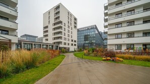 A modern residential building complex with a central paved walkway, surrounded by well-maintained lawns and autumnal foliage. - Warsaw Postępu Apartment - Parking, Balcony, A/C - by Rentujemy (Warszawa)