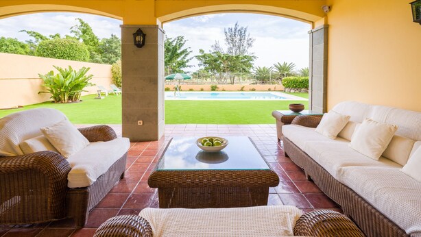 A covered patio with wicker furniture, a glass-top table, and a view of a swimming pool and greenery.