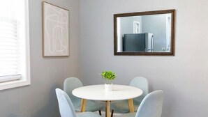 Een eetgedeelte met een ronde tafel, vier lichtblauwe stoelen, een potplant, een lijst met abstracte kunst en een spiegel die een koelkast weerspiegelt.