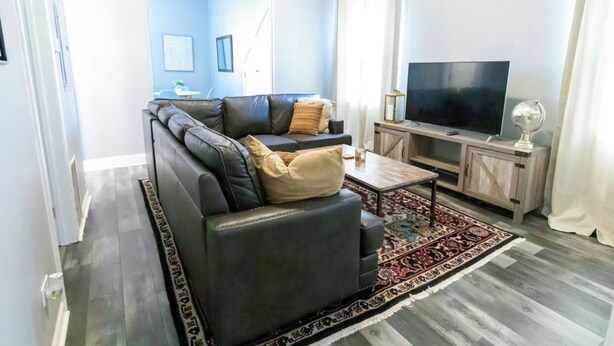 Een woonkamer met een hoekbank, een televisie op een houten kast en een kamerkleed met een patroon.