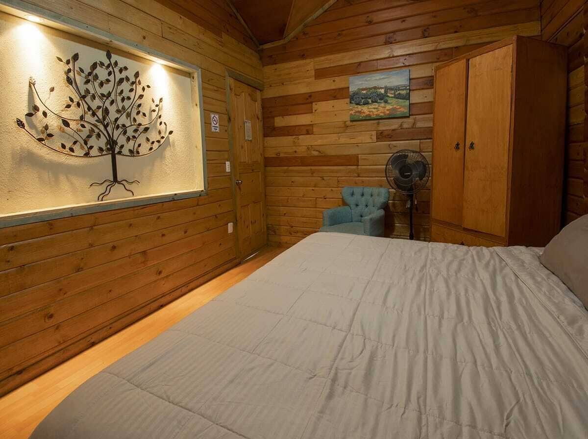 A wooden cabin room with a bed, a chair, a fan, and a wall-mounted artwork.