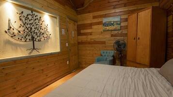 A wooden cabin room with a bed, a chair, a fan, and a wall-mounted artwork.