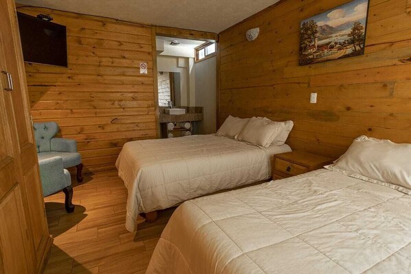A wooden-paneled room with two beds, a chair, a TV, and a painting on the wall.