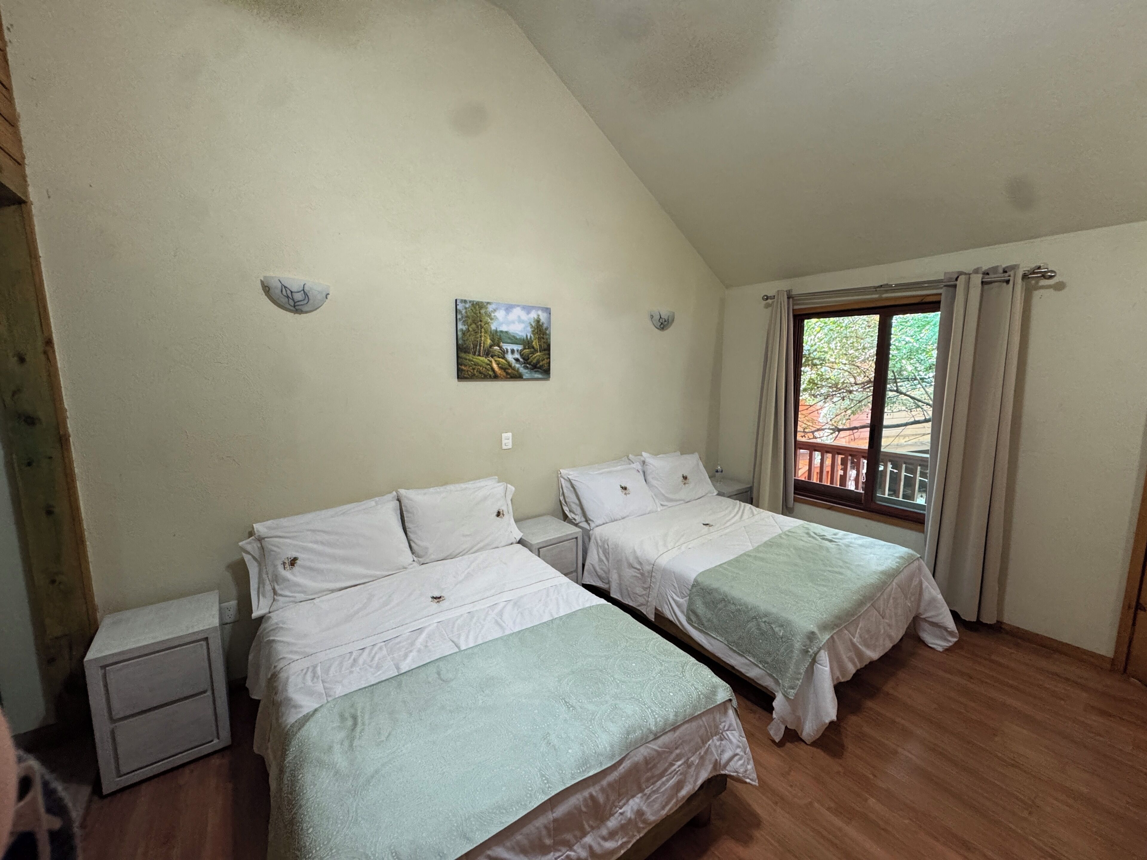 Una habitación con dos camas individuales, una mesita de noche pequeña, un cuadro colgado en la pared y una ventana con cortinas.