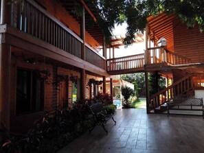 A wooden covered outdoor area with a staircase leading to a second level, surrounded by potted plants and a view of a garden.