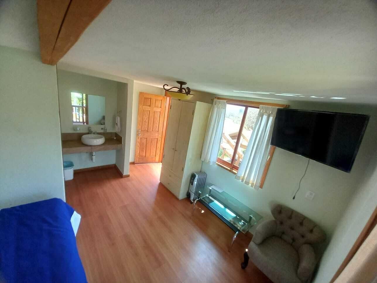A compact room with a wooden floor, a small kitchenette, a bench, and a TV mounted on the wall.
