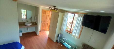 Una habitación compacta con suelo de madera, una kitchenette pequeña, un banco y un televisor montado en la pared.
