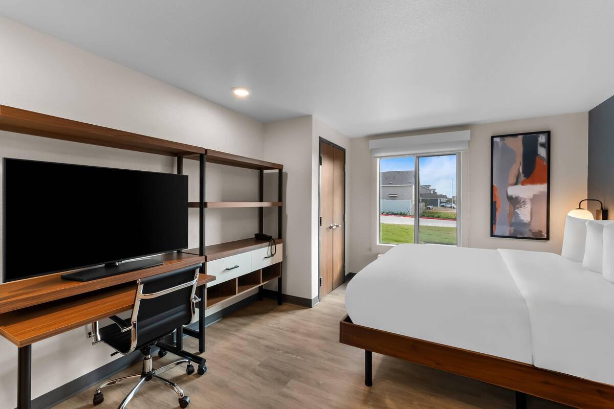 A modern hotel room with a large bed, a desk with a flat-screen TV, a chair, and a window offering a view of the outdoors.