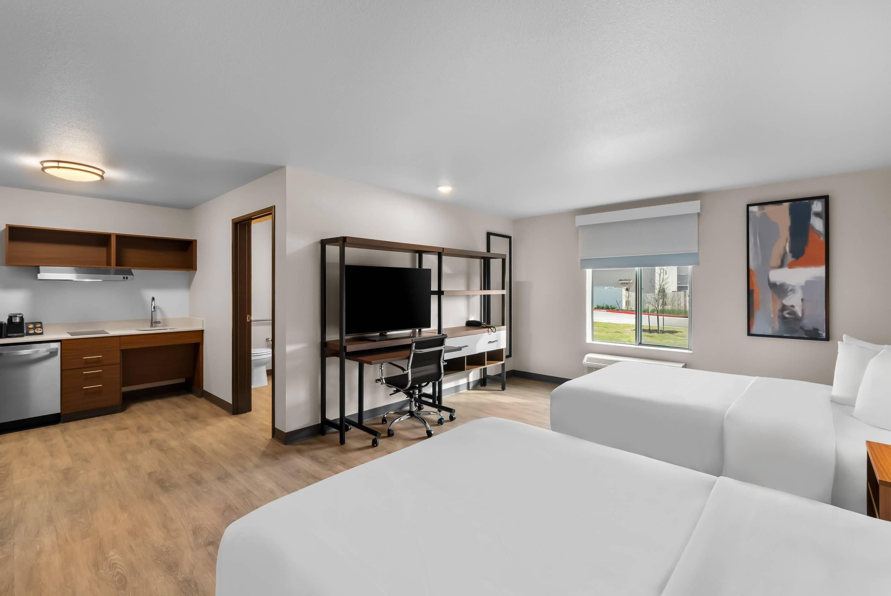Une chambre d’hôtel moderne dotée d’une kitchenette, d’un grand lit, d’un bureau avec une télévision et d’une fenêtre donnant sur la ville.