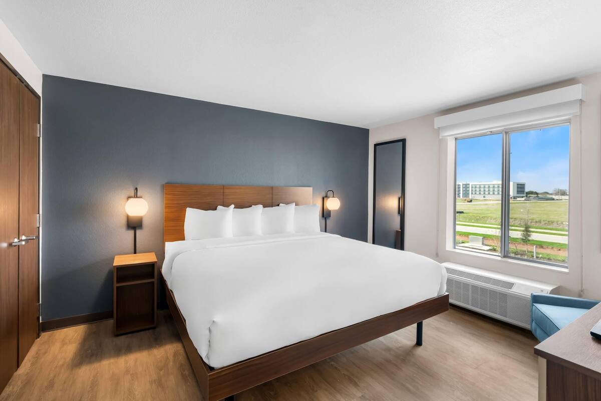 A modern hotel room with a large bed, a nightstand, a desk, and a window with a view of a building and greenery.