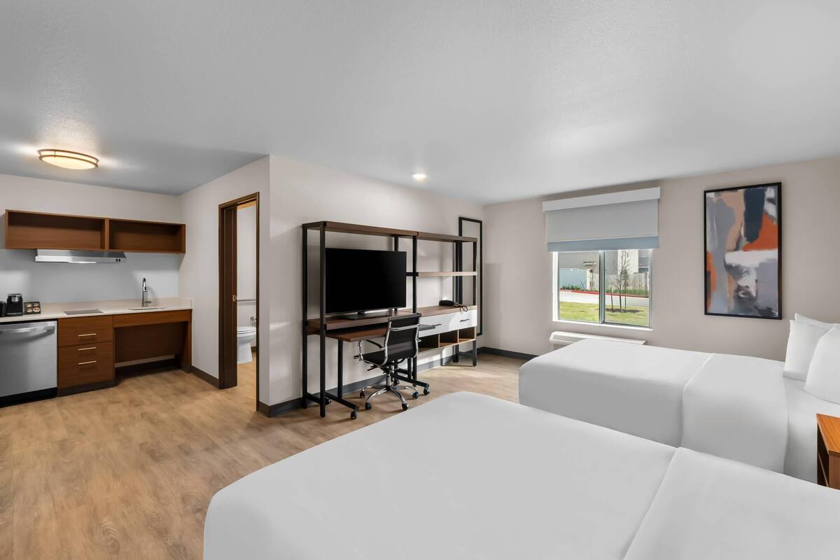 A modern hotel room with a kitchenette, a large bed, a desk with a TV, and a window with a city view.