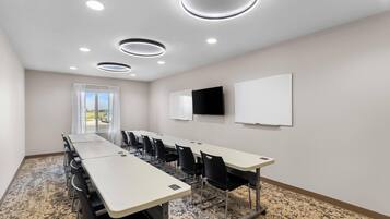 A conference room with a long table, chairs, a whiteboard, a TV, and a window with curtains.