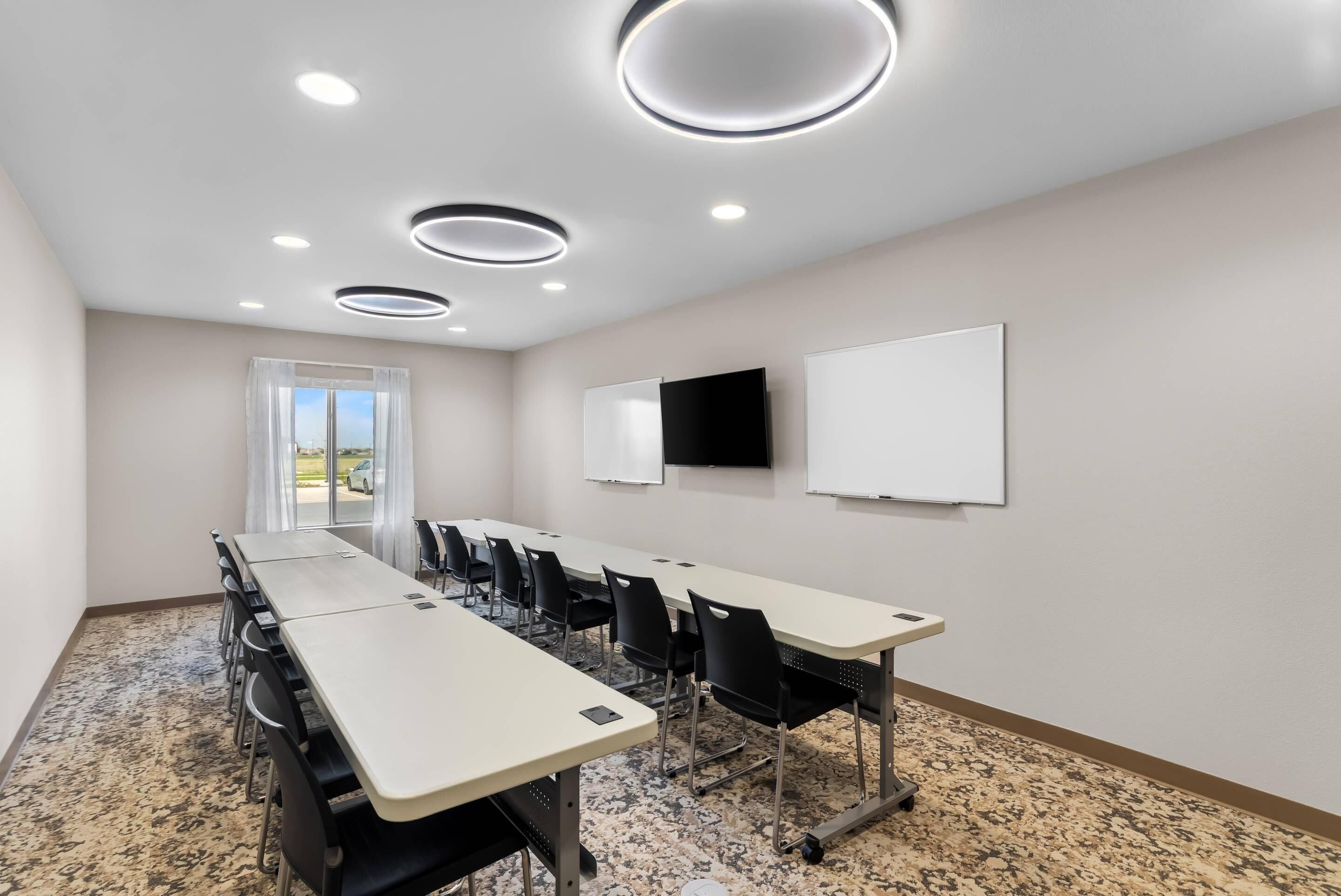 Uma sala de conferências com uma mesa comprida, cadeiras, um quadro branco, uma televisão e uma janela com cortinas.