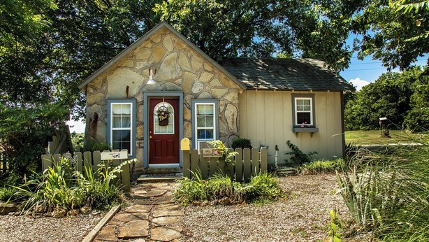 Une petite maison en pierre avec une porte rouge, un petit porche et un chemin de jardin menant à l’entrée.