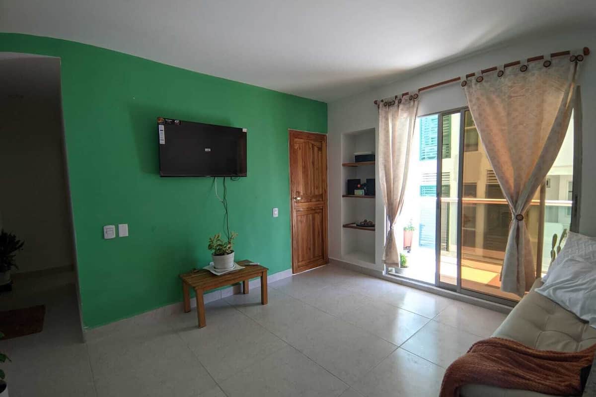 Un salon avec un mur vert, un téléviseur à écran plat fixé au-dessus d’une table en bois sur laquelle est posée une plante en pot, une porte-fenêtre coulissante offrant une vue dégagée et un canapé beige.