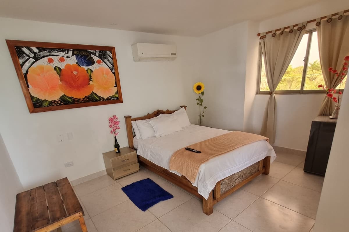 Une chambre à coucher comprenant un lit, une table de chevet, une petite table, un tableau, un climatiseur, une fenêtre avec des rideaux et un vase avec des fleurs.