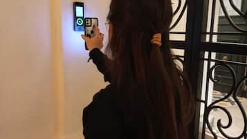 A person taking a photo with a smartphone near a wall-mounted device, possibly a security camera or intercom, in a room with a black metal gate or door.