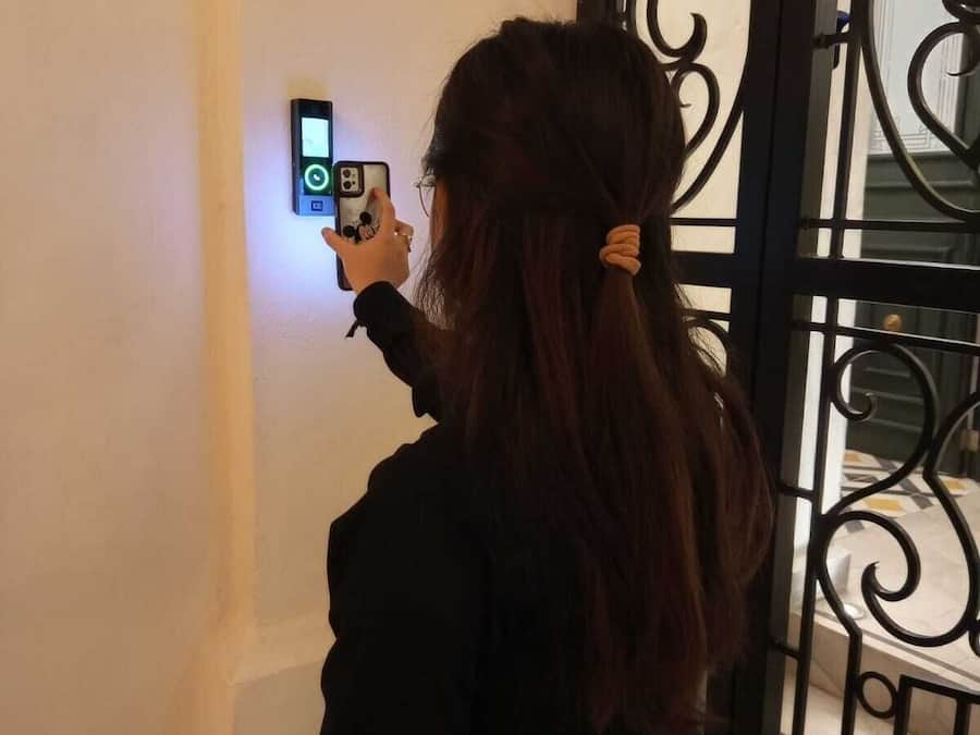 A person taking a photo with a smartphone near a wall-mounted device, possibly a security camera or intercom, in a room with a black metal gate or door.