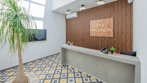 A hotel reception area with a modern design, featuring a wooden panel wall, a reception desk, and a large potted plant. - Hotel Miranda (Querétaro)