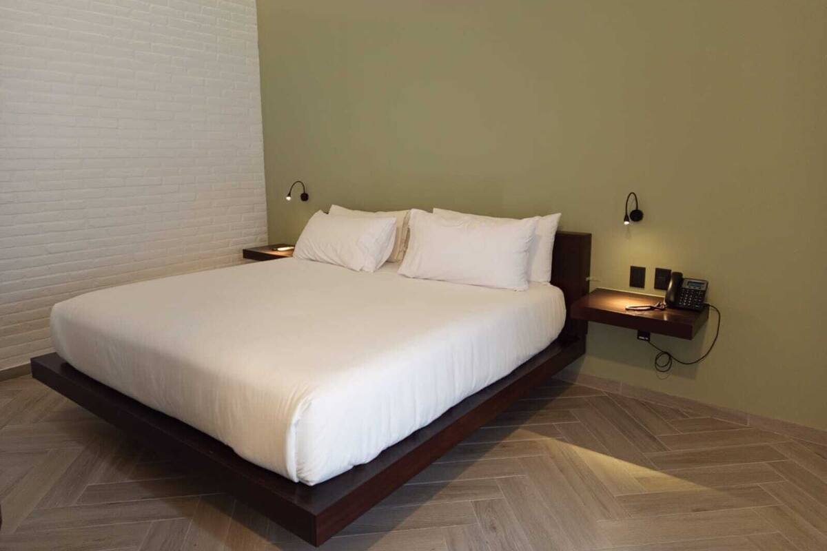 A neatly made bed in a room with a herringbone floor pattern, a bedside table with a phone, and a wall-mounted light fixture.
