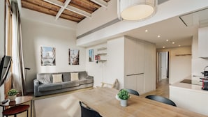 Een moderne woonkamer met een bank, een eettafel en stoelen. Er is een keuken met een spoelbak en kastjes.
