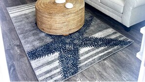 Una cocina moderna con armarios blancos, una zona de comedor con mesa y taburetes blancos, un sofá gris con cojines estampados y un tapiz de rayas azul y gris.