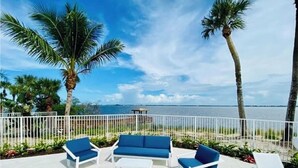 Un área de estar al aire libre con mobiliario blanco y azul, palmeras y vista al mar.