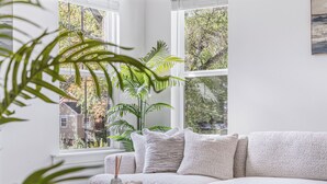 A modern living room with a white sofa, a round table, and a large window with a view of trees and buildings. - Modern Home Near Uptown & Camp North End - Sleeps 12, Sports-Themed Game Room (Charlotte)