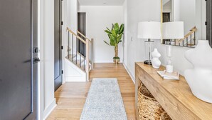 A hallway with a wooden floor, a light blue rug, a wooden console table with a lamp, a large mirror, and a potted plant. - Modern Home Near Uptown & Camp North End - Sleeps 12, Sports-Themed Game Room (Charlotte)