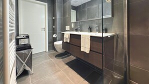 Un bagno moderno con una parete rivestita in piastrelle scure, una porta bianca, una cabina doccia in vetro e un mobile lavabo con due lavandini.