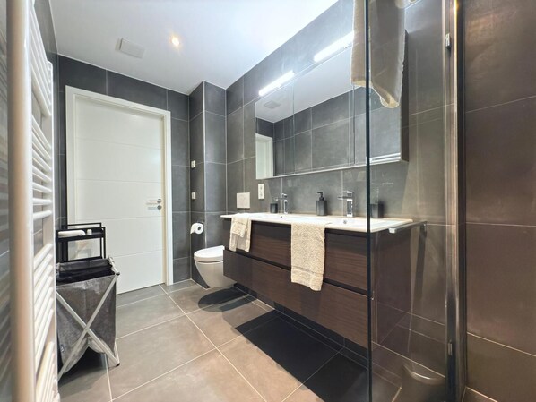 Un bagno moderno con una parete rivestita in piastrelle scure, una porta bianca, una cabina doccia in vetro e un mobile lavabo con due lavandini.