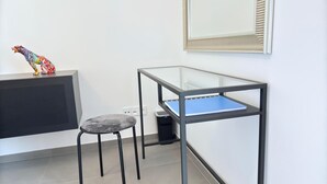 Una stanza moderna e accogliente con un tavolino in vetro, uno sgabello imbottito grigio, una televisione e uno specchio a parete.