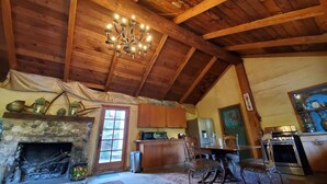 A rustic interior with a stone fireplace, wooden ceiling beams, a chandelier, and a dining area with a table and chairs.