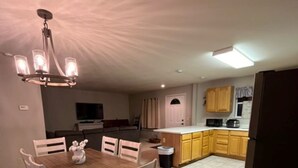 A dining area with a wooden table and chairs, a chandelier, and a kitchen with yellow cabinets and a refrigerator.