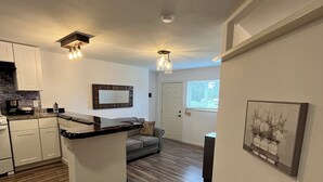 A modern kitchen with white cabinets, a black countertop, and a herringbone wood floor. There is a gray sofa in the living area and a framed picture on the wall.