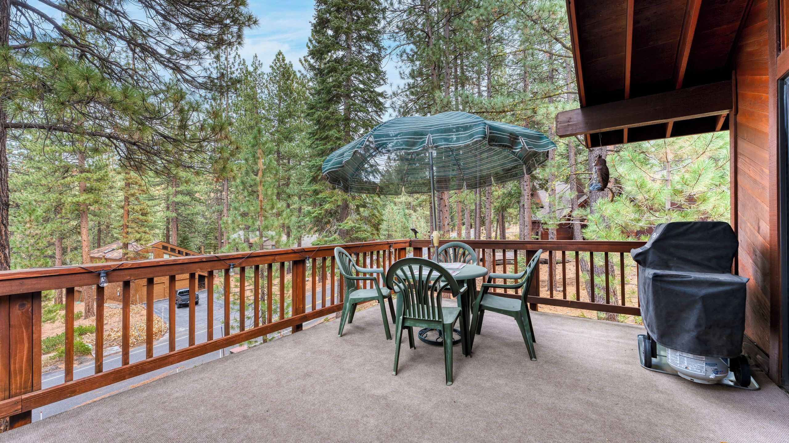 A covered outdoor deck with a table and chairs, surrounded by a wooden railing, overlooking a forested area.