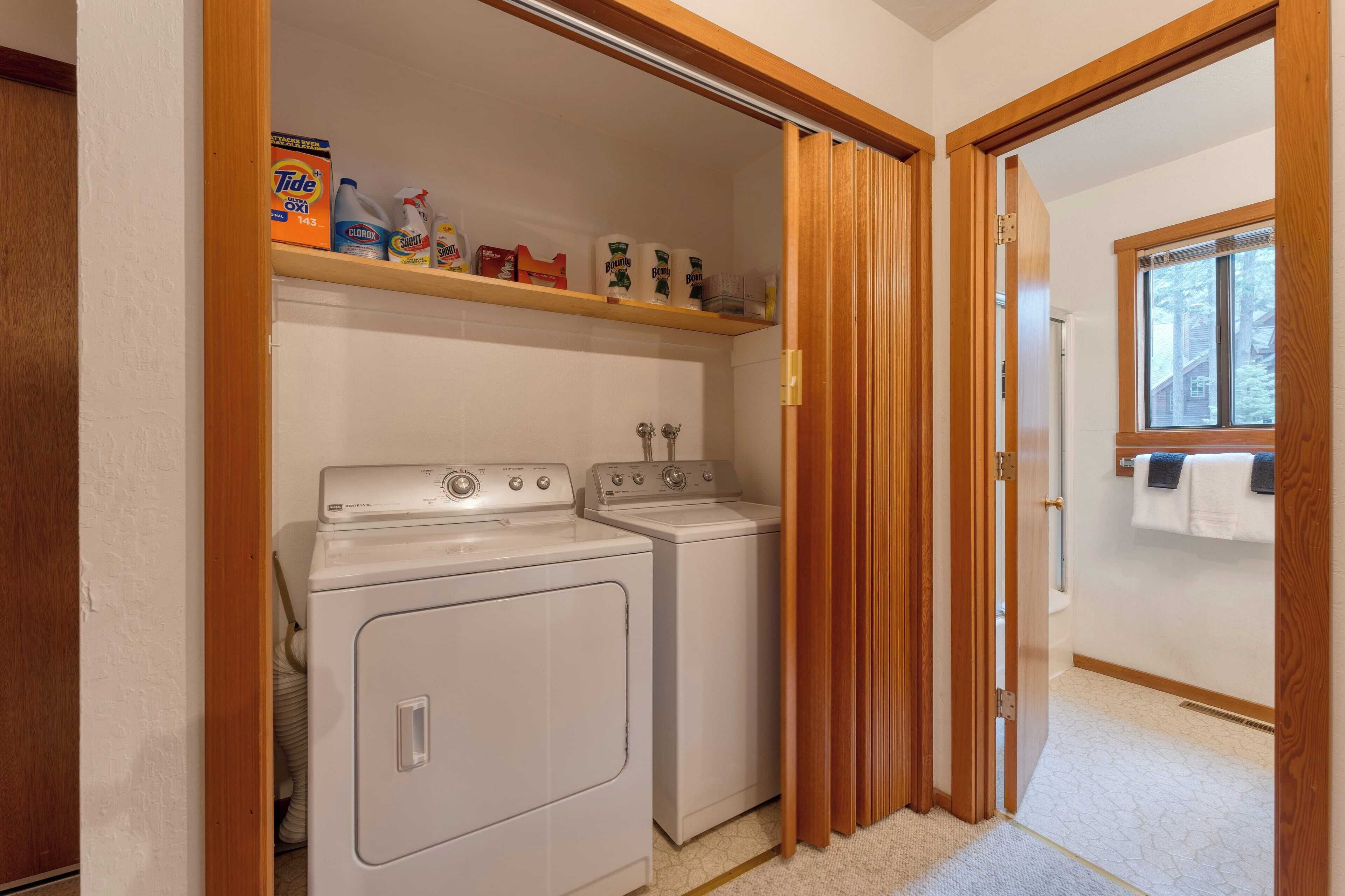 A laundry room with two white washers and dryers, a shelf with cleaning supplies, and a window with towels hanging on the wall.