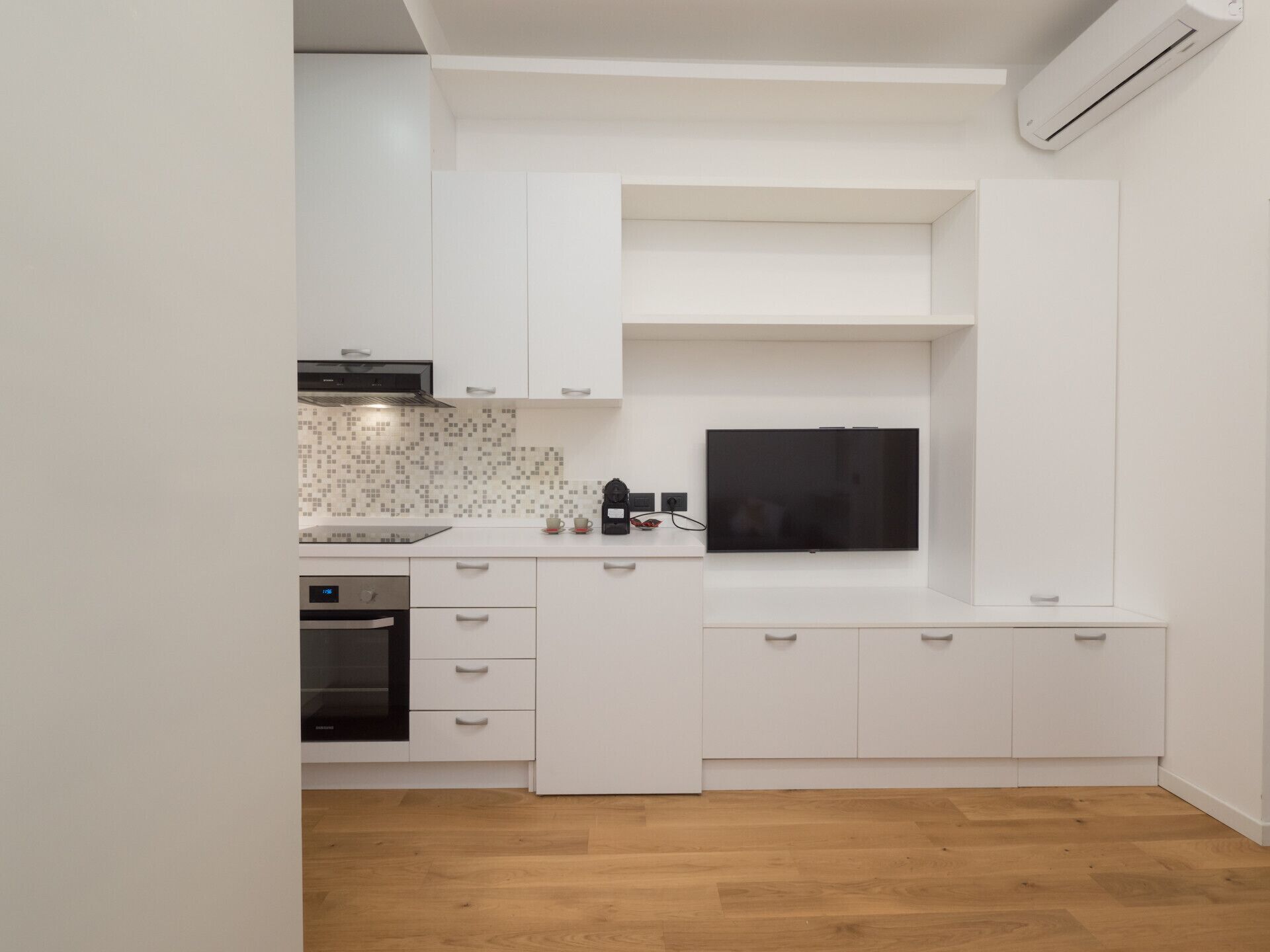 A modern kitchen with white cabinetry, a built-in oven, and a flat-screen TV mounted above the countertop.