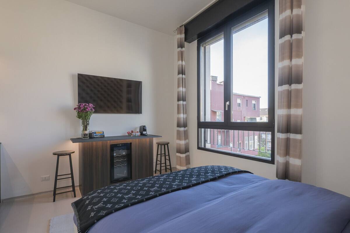 A modern hotel room with a large bed, a small desk with a chair, a television, and a window with curtains.