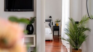 A modern hotel room with a large flat-screen TV, a bed with white bedding, a bedside table with a lamp, and a potted plant. - Corso Garibaldi - Modern & Cozy Suite by Rentopolis (Milan)