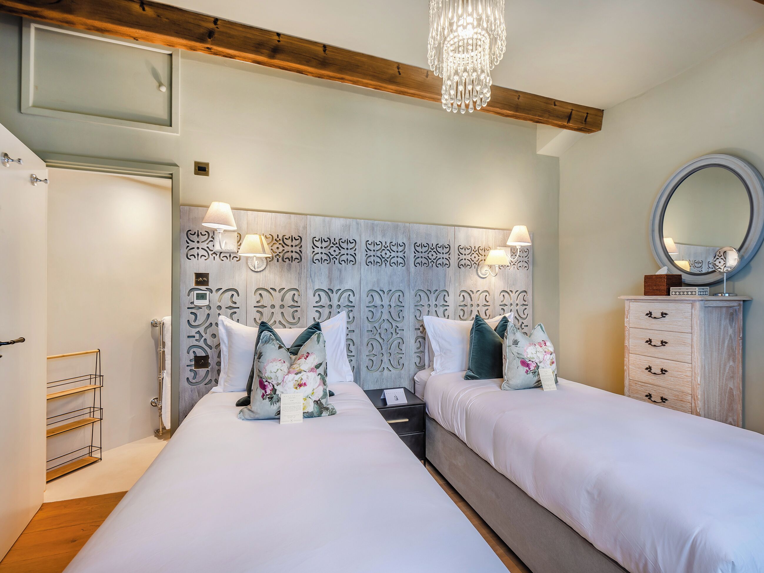 A hotel room with two beds, a wooden ceiling, a chandelier, a decorative headboard, bedside lamps, a mirror, and a wooden dresser.