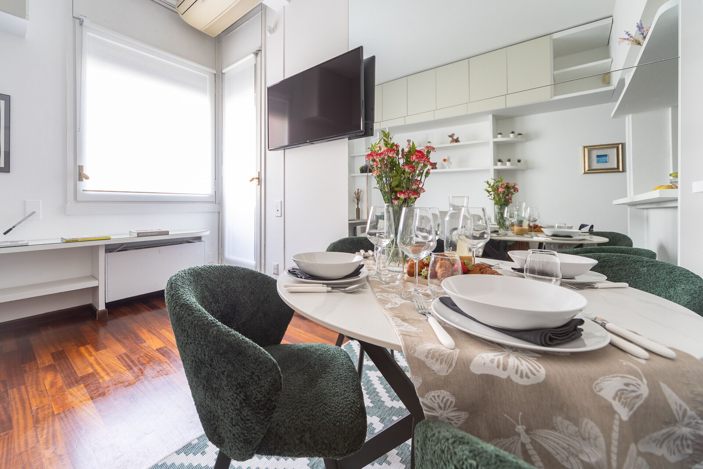 A modern dining room with a round table set for four, green upholstered chairs, a flat-screen TV mounted on the wall, and a window providing natural light.